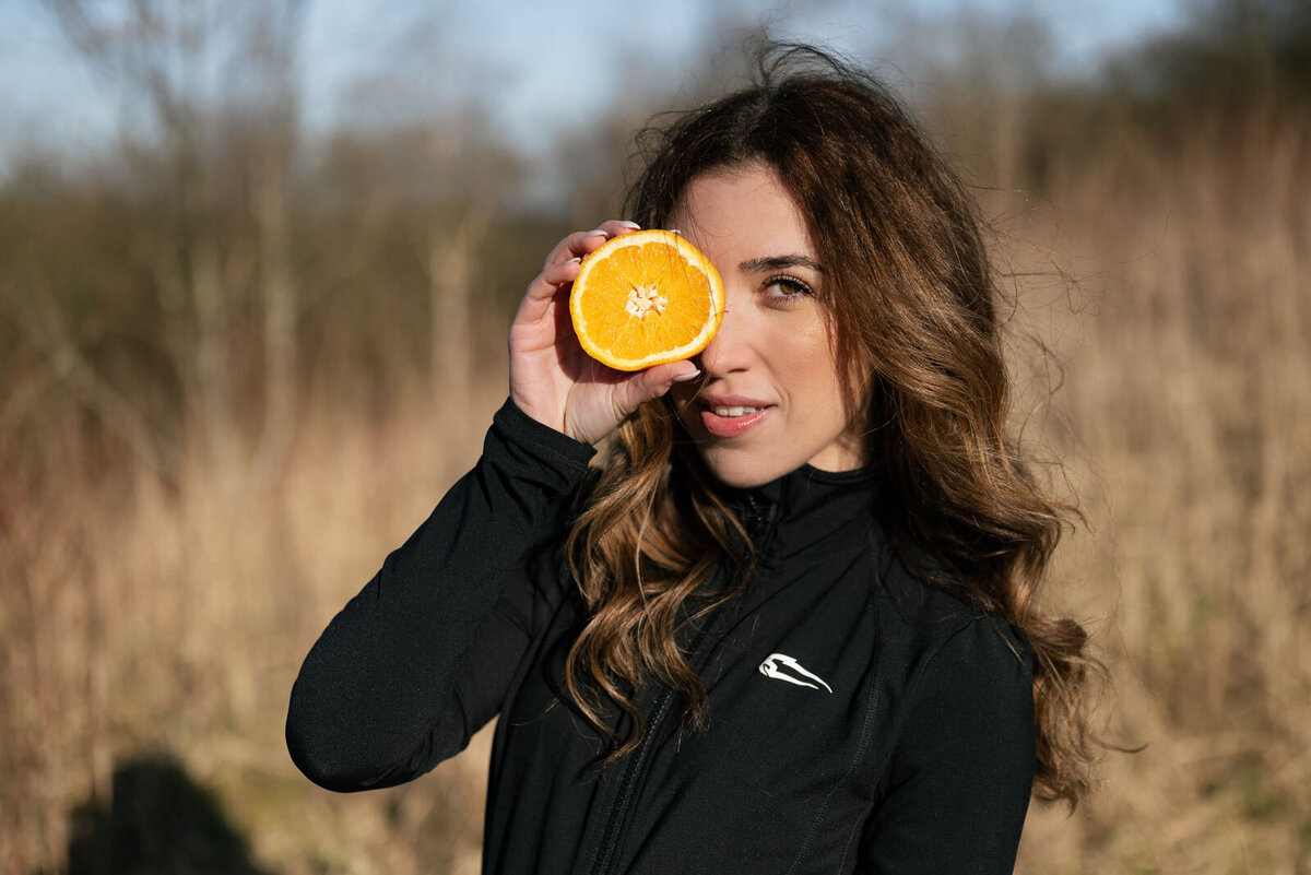 eine Ernährungsberaterin steht im Feld in ihrer Sportkleidung und hält eine halbe Orange vor ihr rechtes Auge