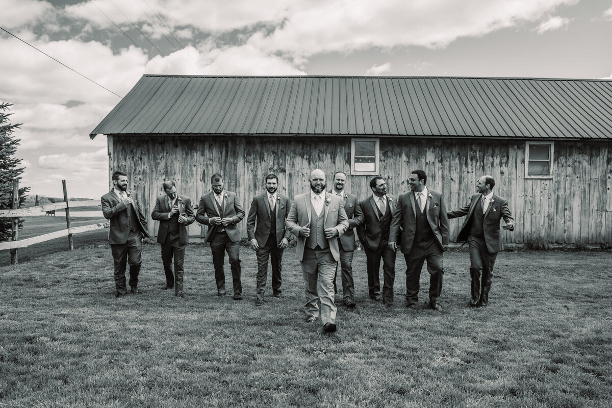 groom and groomsman walk in front of old barn in black and white
