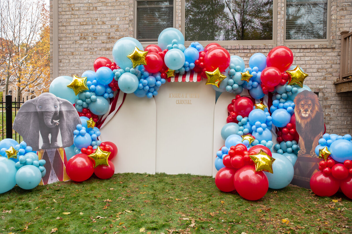 Cardboard circus animals and red, blue, and gold balloons set up on white wooden arches.