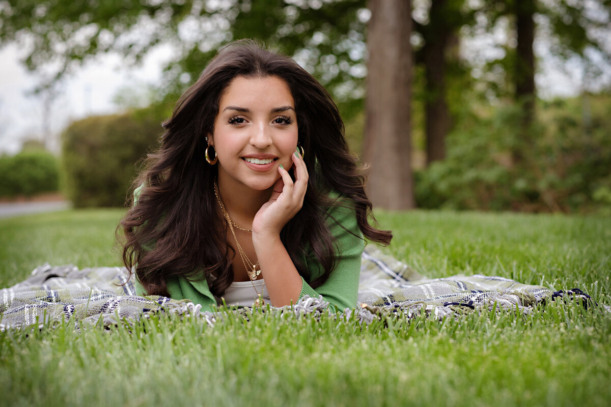High school senior lying on a plaid blanket in the grass wearing a green cardigan and gray jeans, captured in a relaxed spring portrait.