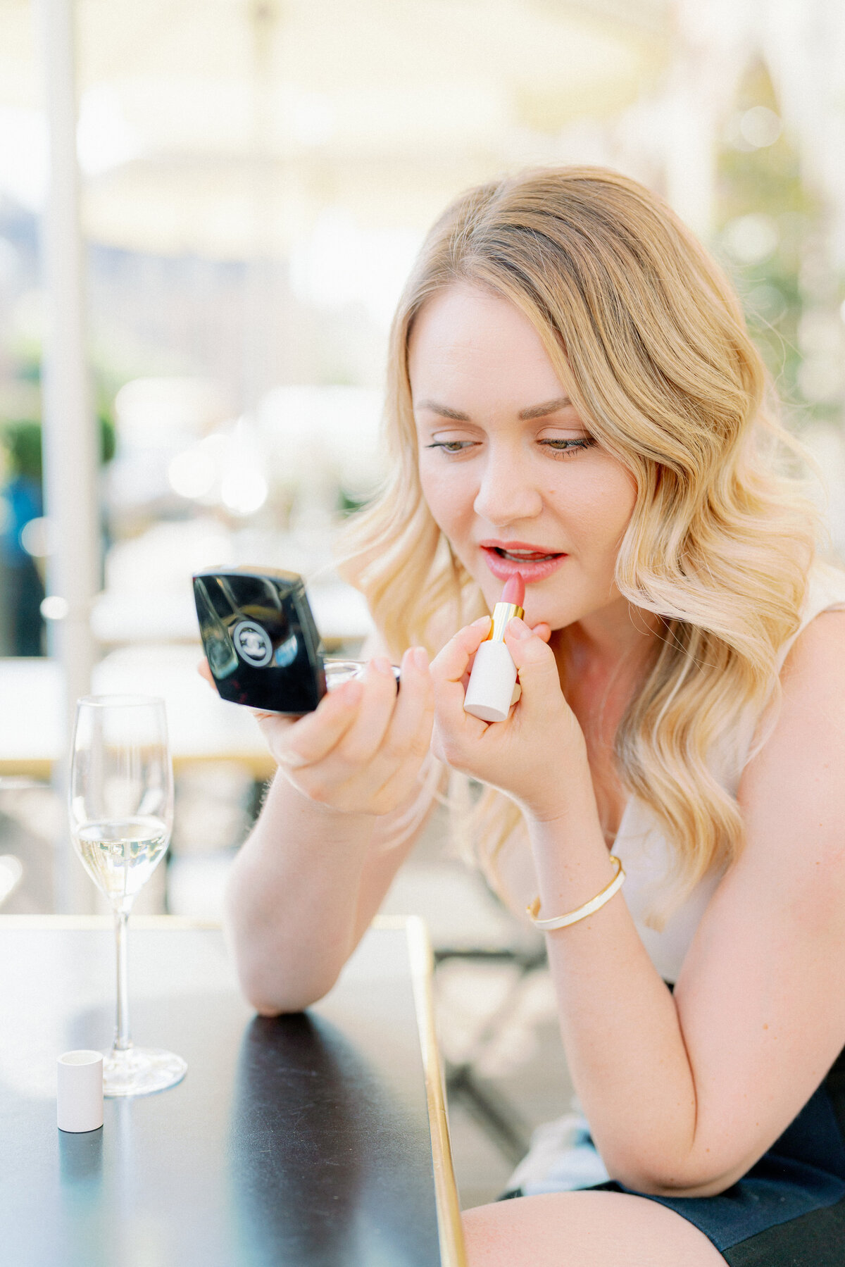 a blonde woman putting lipstick on at a table at a cafe in paris france