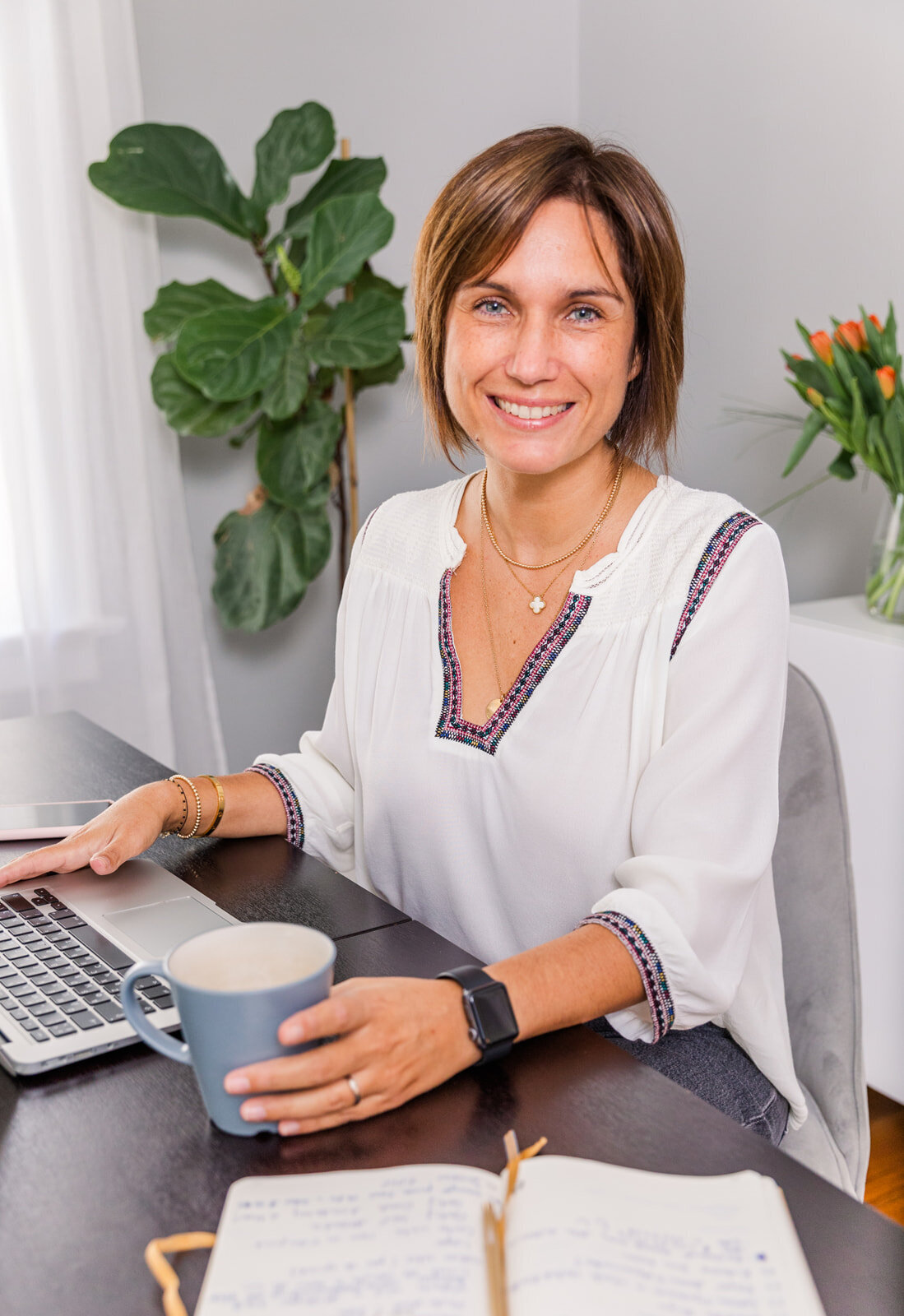 coach wearing white top working at her desk with her laptop, coffee and notebook