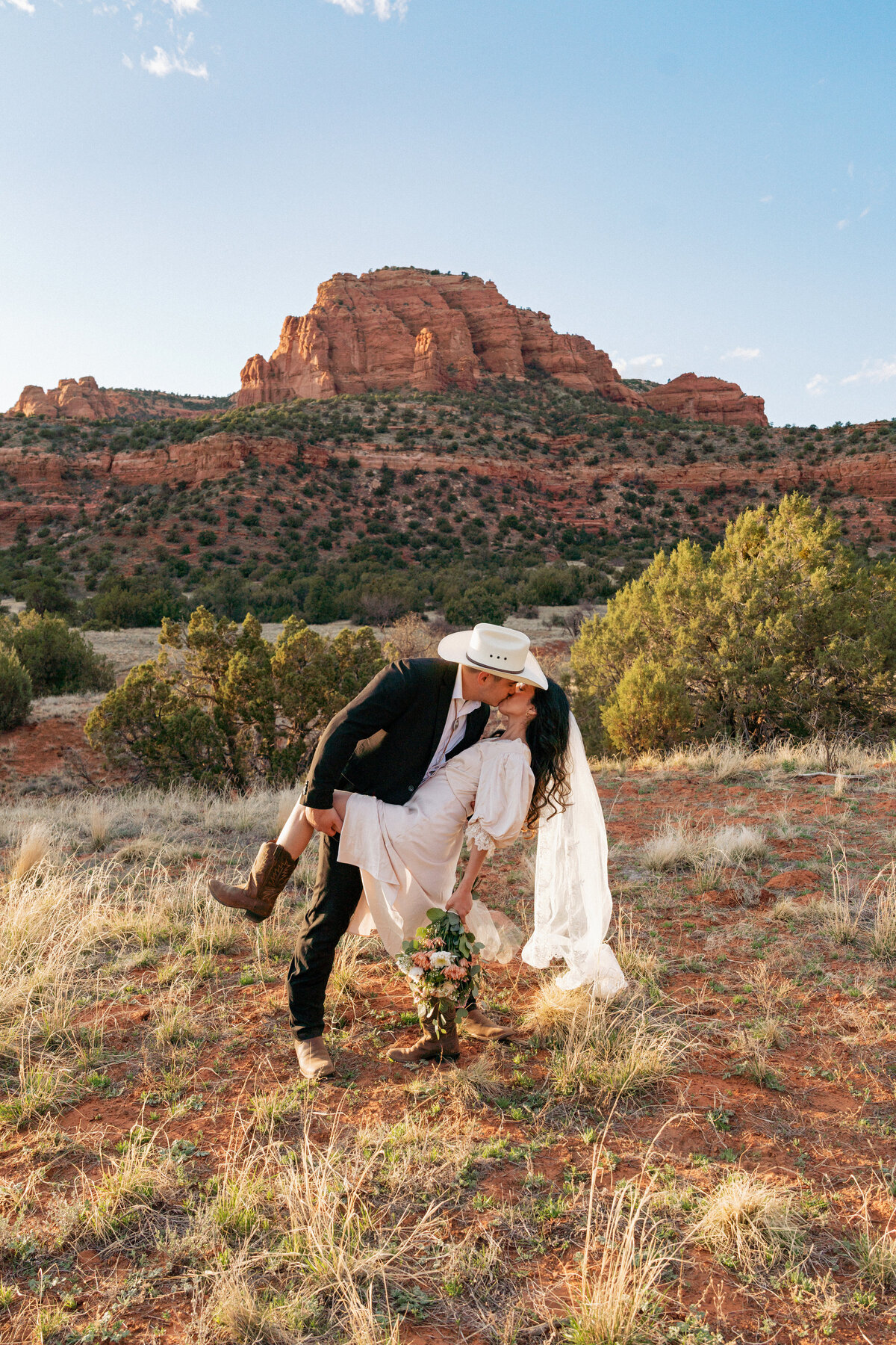 Groom in cowboy hat kisses his bride during a western-inspired elopement at Bear Mountain in Sedona, Arizona, surrounded by red rock formations and desert landscape.