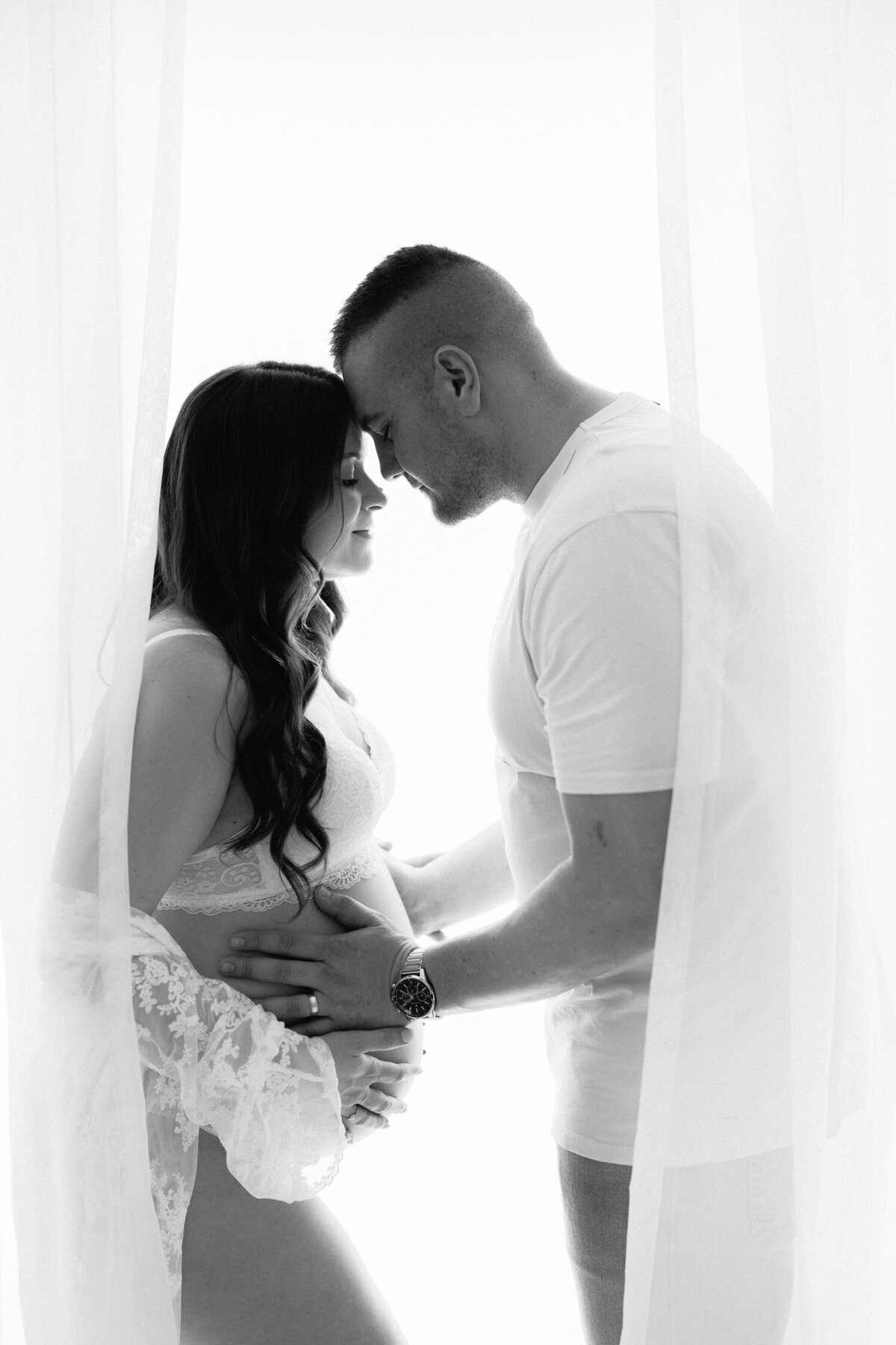 Black and white image of pregnant woman posing in white studio with partner holding her belly gently, with their foreheads touching together.