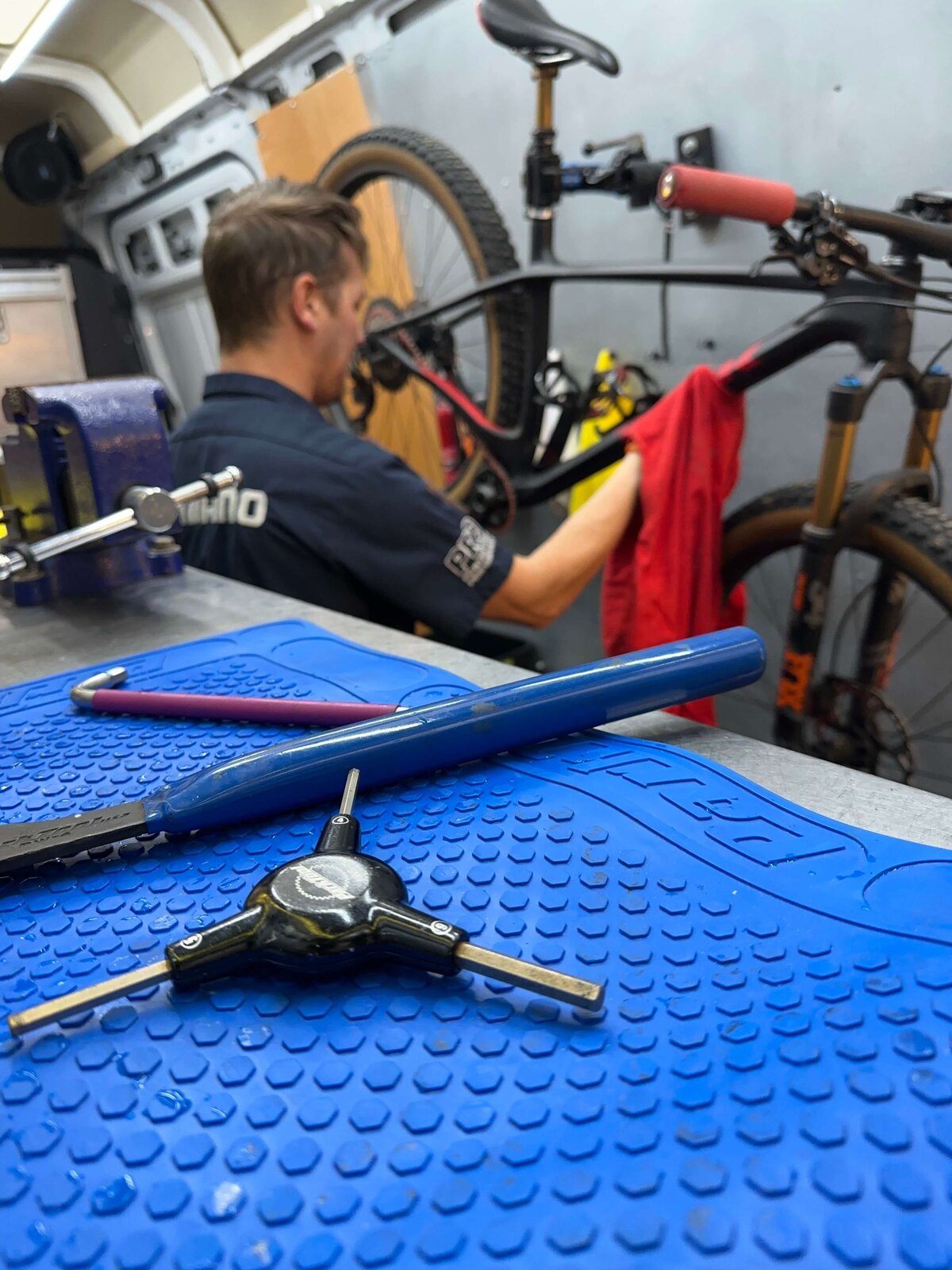 A bicycle mechanic wearing a Shimano shirt works on a mountain bike, wiping it with a red cloth. A blue mat with tools, including a hex wrench and screwdriver, is in the foreground