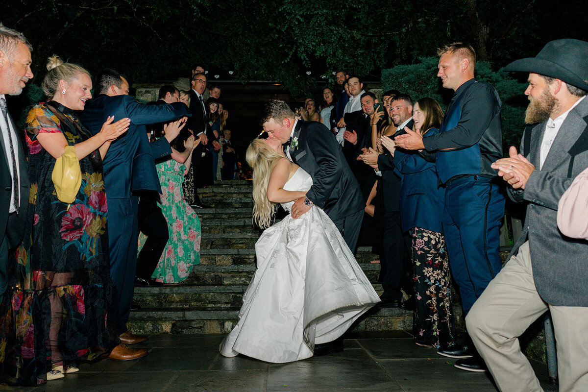  Bride and groom share a dramatic kiss during their wedding exit, surrounded by cheering guests lined along the stone steps at Old Edwards Inn in Highlands, North Carolina.