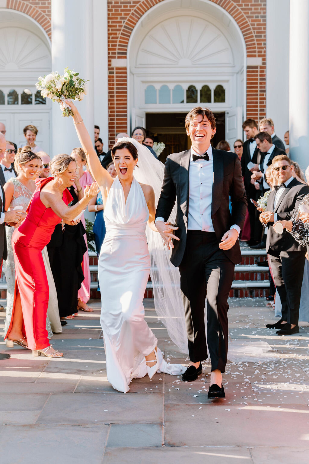Rhode Island Wedding Photographers | A joyful bride and groom celebrate with guests as they leave a building. The bride lifts her bouquet excitedly, surrounded by applauding friends.