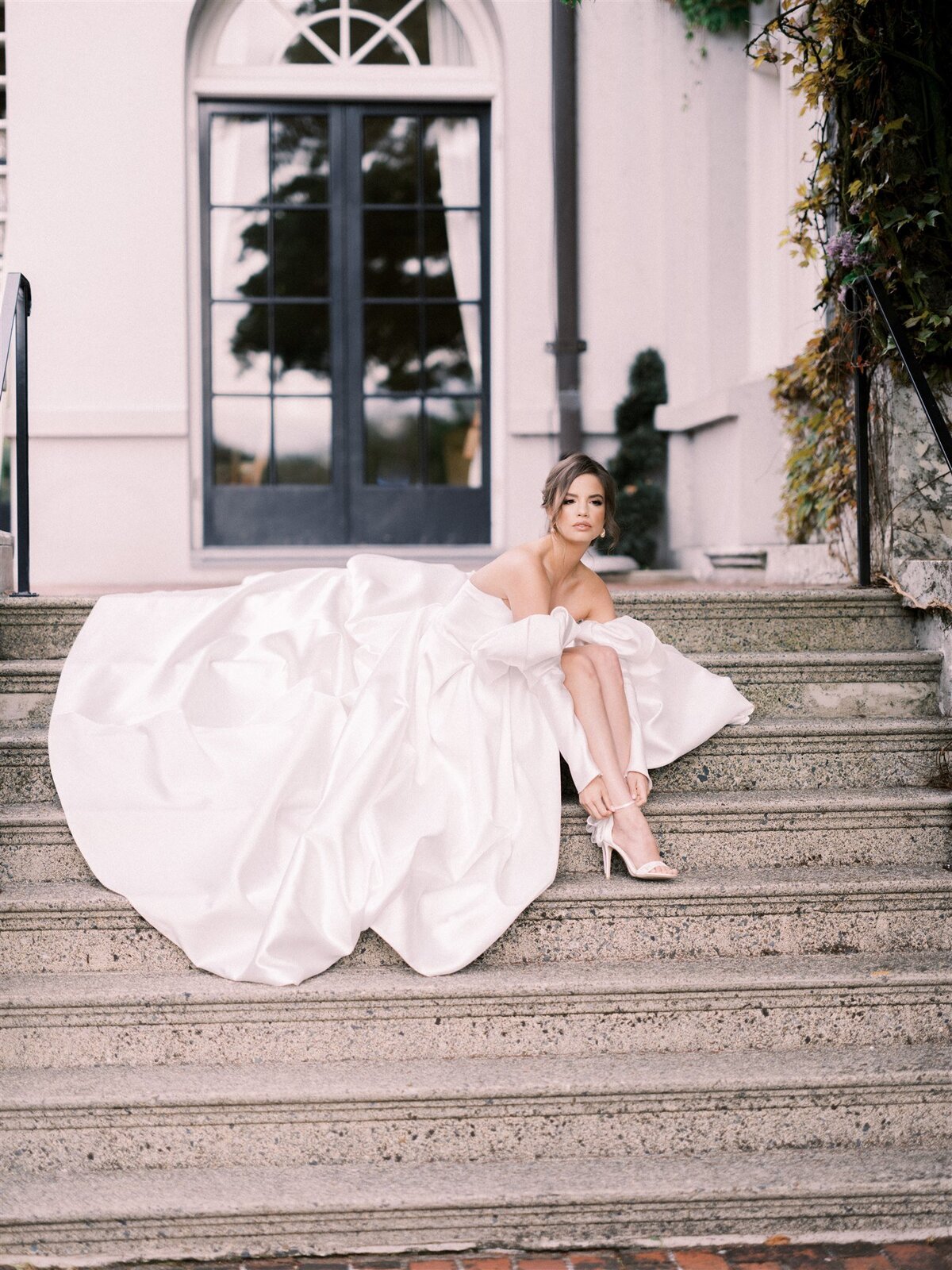 A woman in an off-the-shoulder white wedding dress sits on outdoor stone steps in front of glass doors, looking to her left with one leg extended and wearing white heels. The elegance of the setting reflects the meticulous touch of Destination Wedding Planner Melissa Dawn Event Designs.