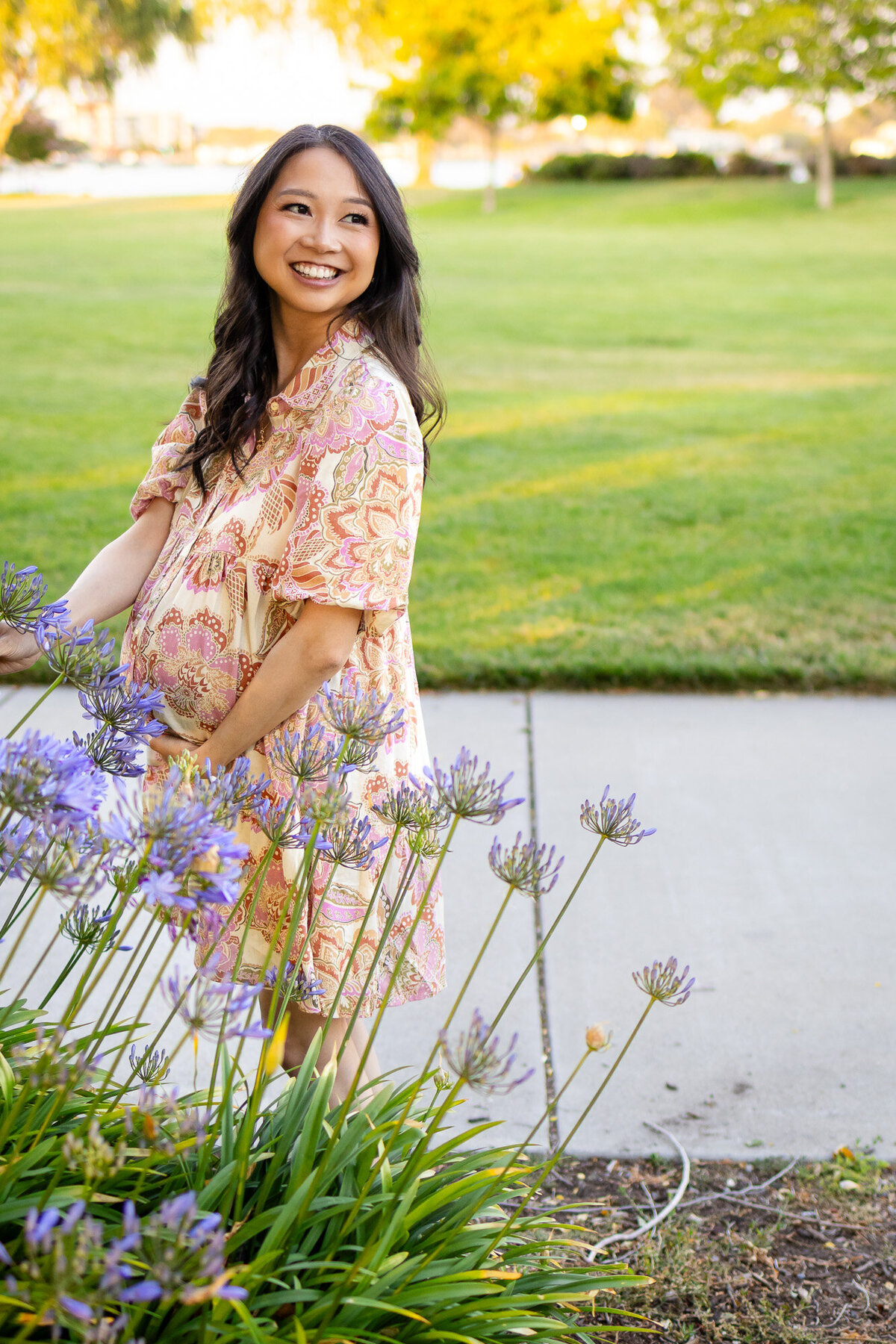 Glowing mother-to-be smiles while holding her belly in a garden filled with blooming purple flowers — Bay Area Maternity Portfolio - Ellobelle Photography.