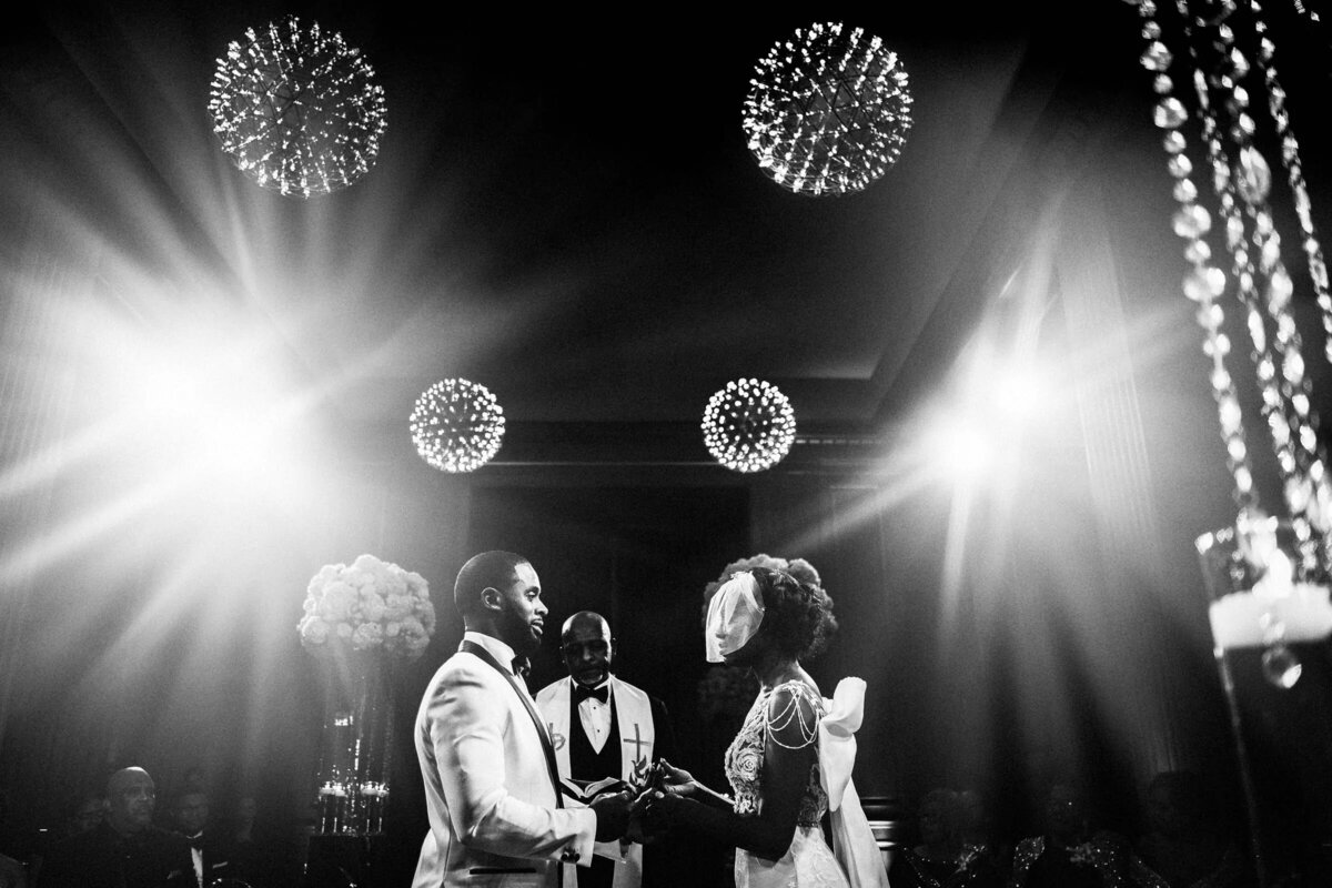 Black and white photo of a bride and groom exchanging vows, with the focus on their hands and the officiant, under a spotlight.