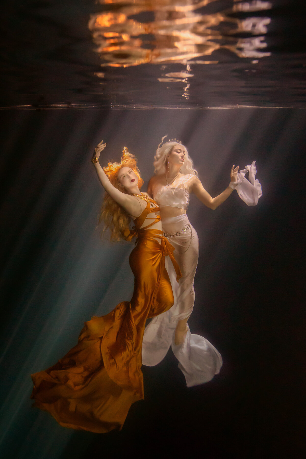 Two women gracefully floating underwater, bathed in soft golden light. One wears a flowing golden gown with intricate detailing, while the other is dressed in elegant white, adorned with delicate accessories. Their movements resemble a synchronized dance, captured in a serene and otherworldly underwater environment.