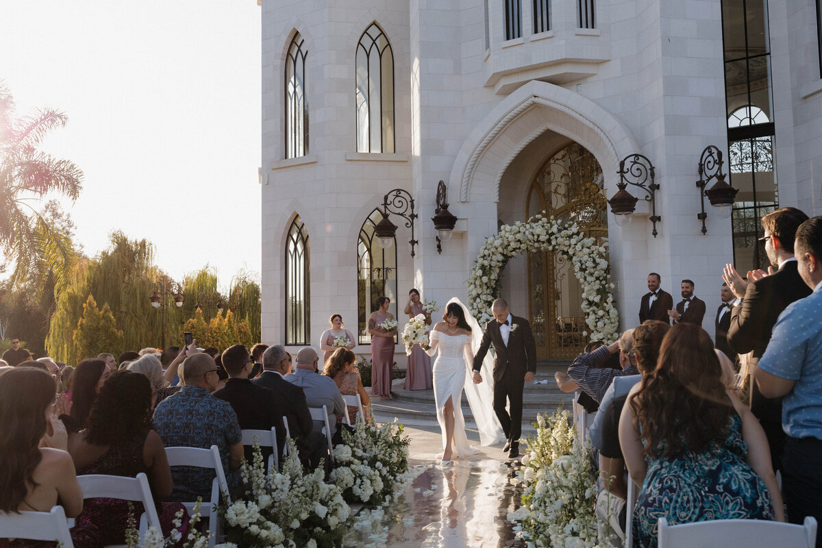 A newlywed couple exiting their wedding ceremony, holding hands and smiling as guests cheer.