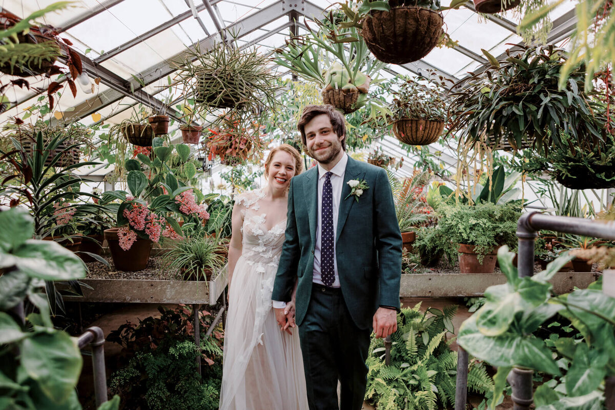The bride and groom are standing inside a greenhouse, with potted plants hanging above them and also around them as well.