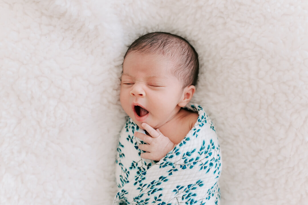 A sleepy newborn girl swaddled in a blue floral pattern yawning