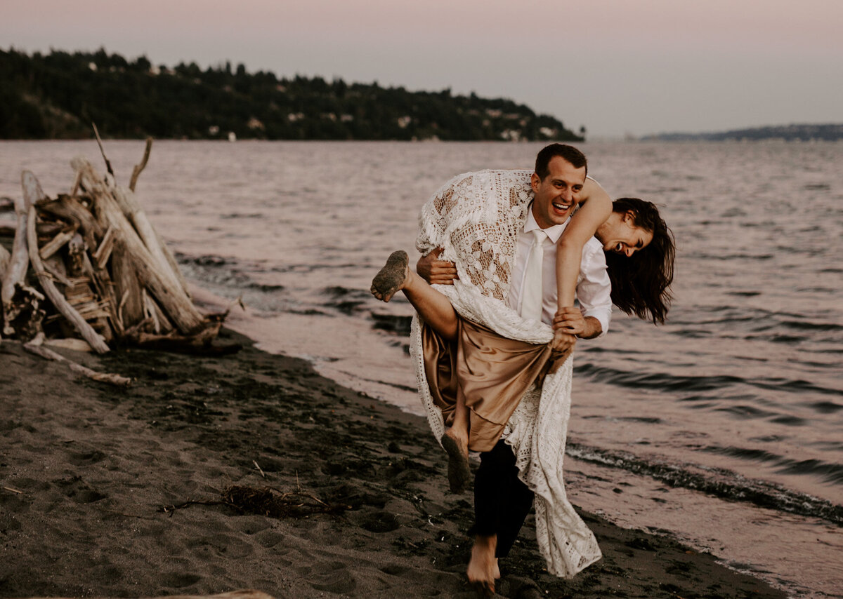Groom carries his bride over his shoulders along a beach in Seattle