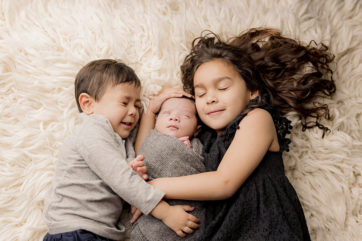 siblings holding baby brother laying down with eyes closed
