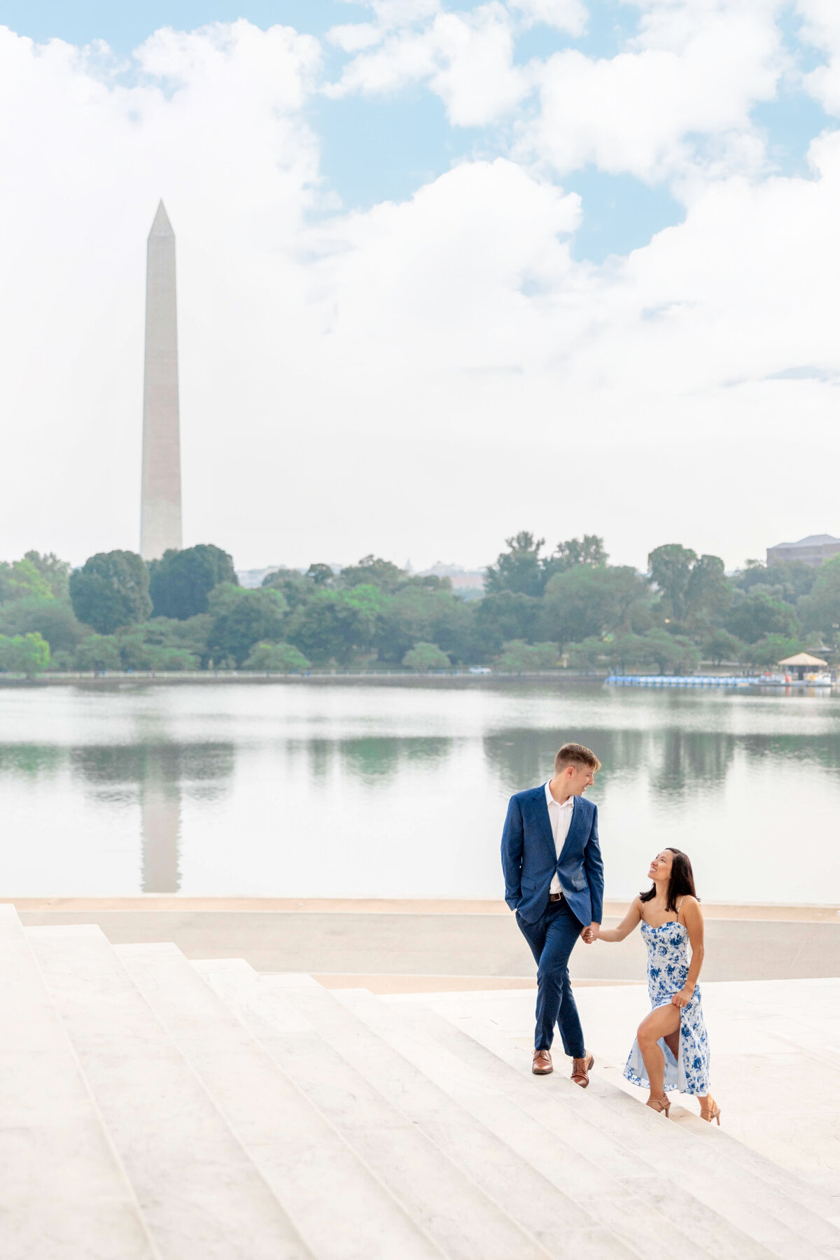 A couple in elegant attire sit on steps by a large reflecting pool, with the Washington Monument in the background. The woman in a floral dress smiles up at the man in a blue suit, who looks down at her, against a backdrop of green trees and a cloudy sky.
