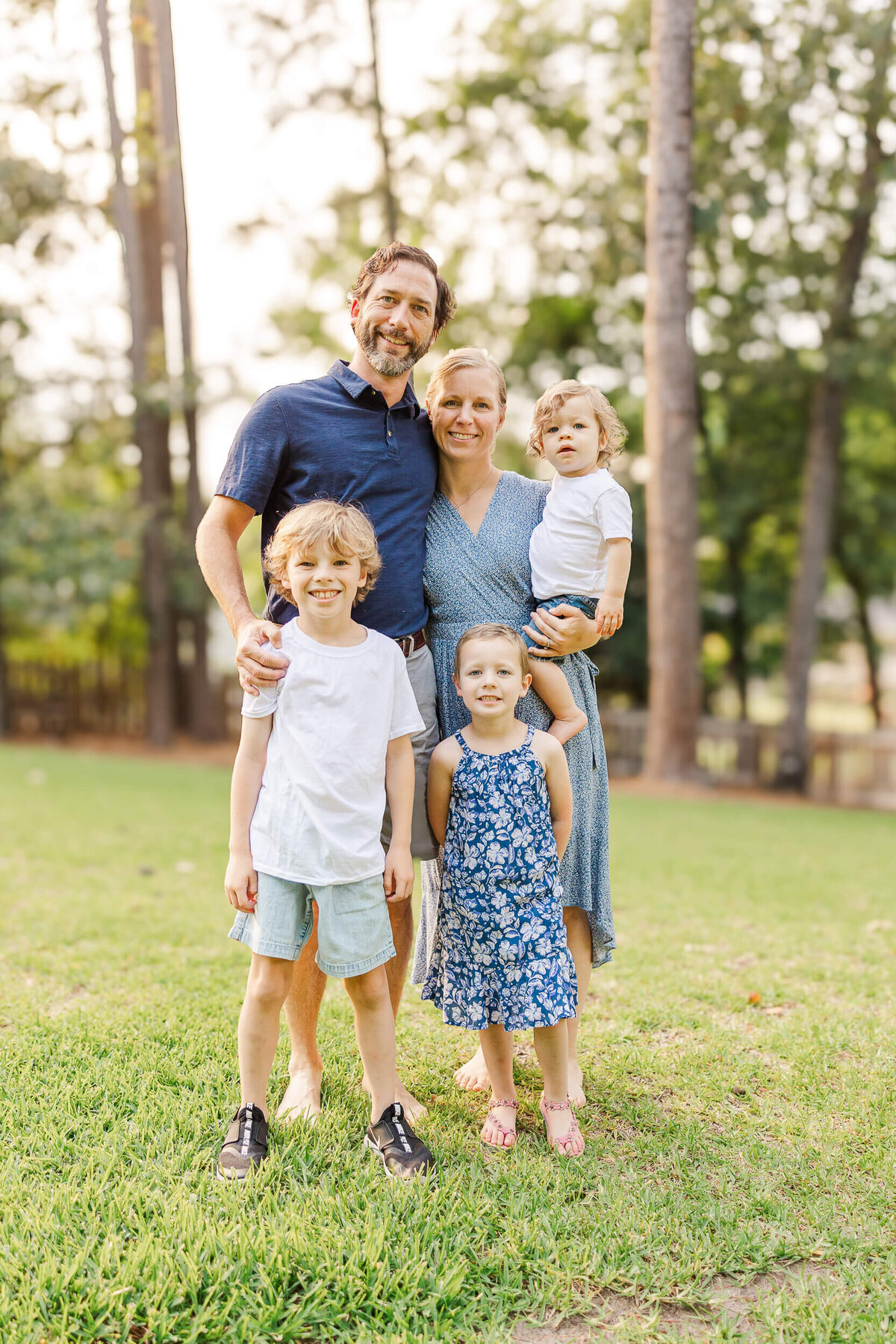 A Fayetteville NC family photographer captures a joyful moment as a family of five stands outdoors on grass with trees in the background. The father, mother, two young children, and a toddler are all smiles. Father and son wear shorts, while mother and daughter don dresses, as the mother holds the toddler.