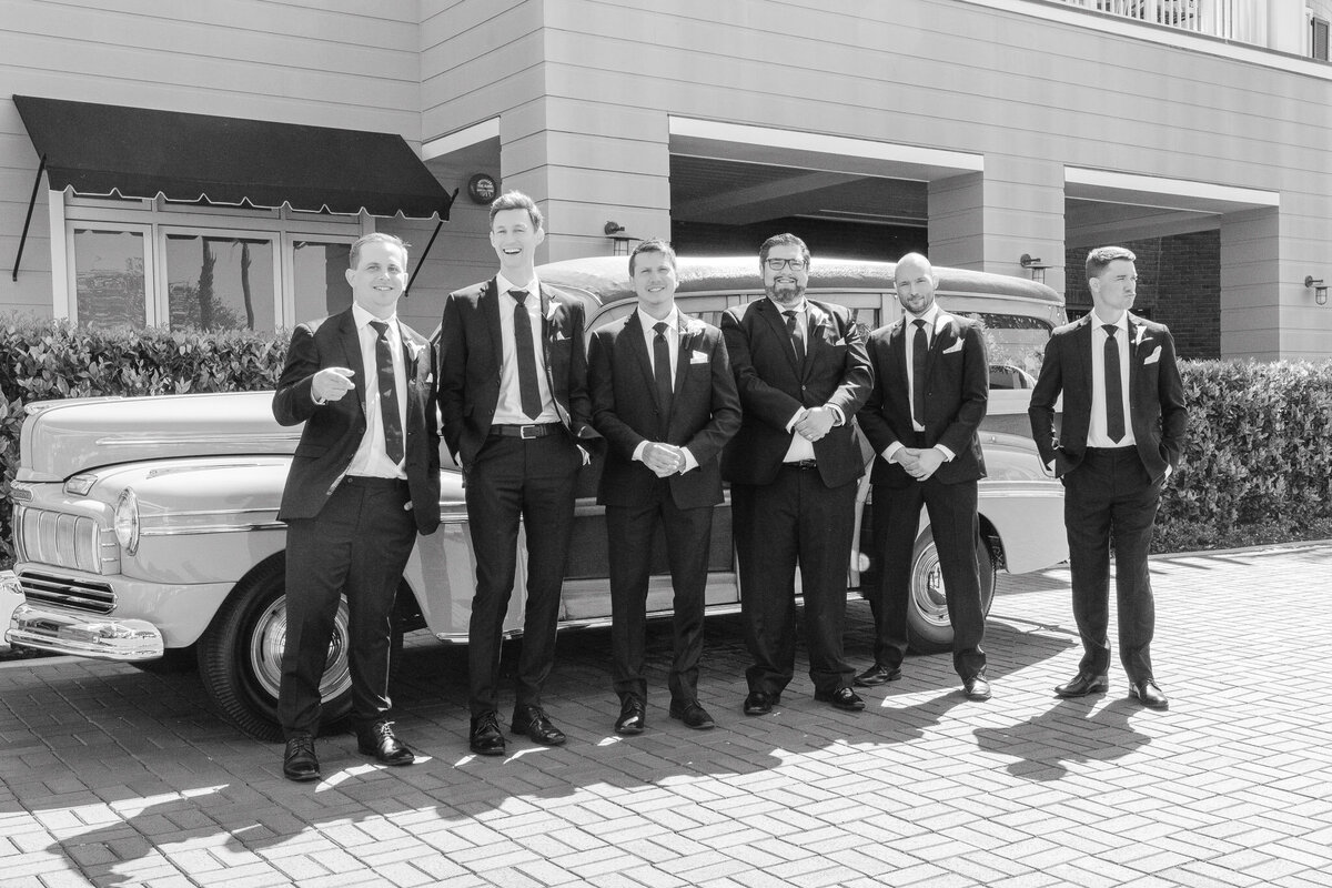 Groom and groomsmen in black suits posing in front of a vintage car at Lido House, Newport Beach