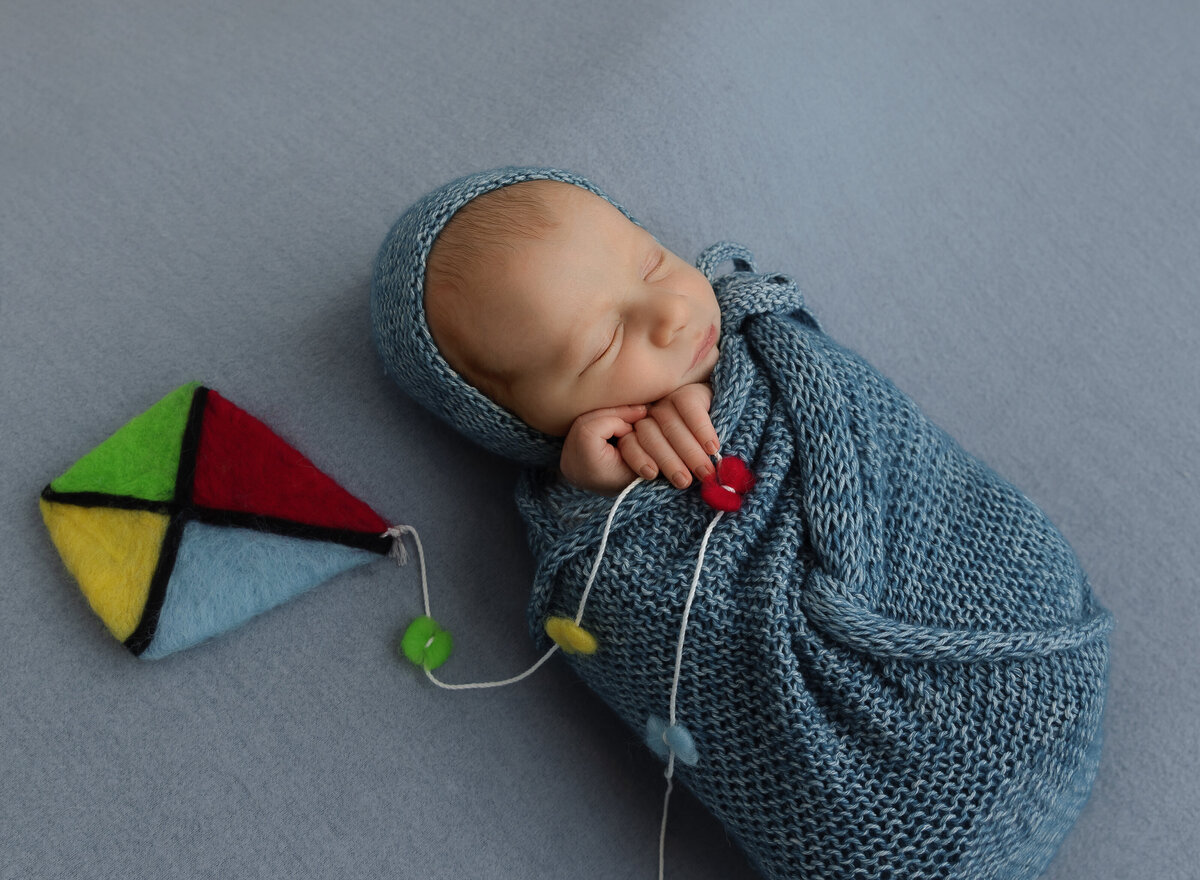 Baby boy swaddled in blue knit is posed for his newborn photoshoot. His hands are peeking out of the swaddle and holding a felt kite. Captured by top Brooklyn newborn photographer Chaya Bornstein