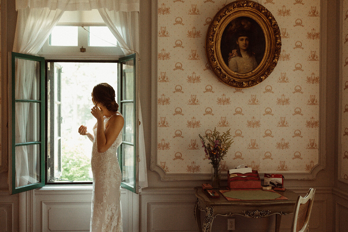 A bride stands in a window while getting ready on her wedding day.
