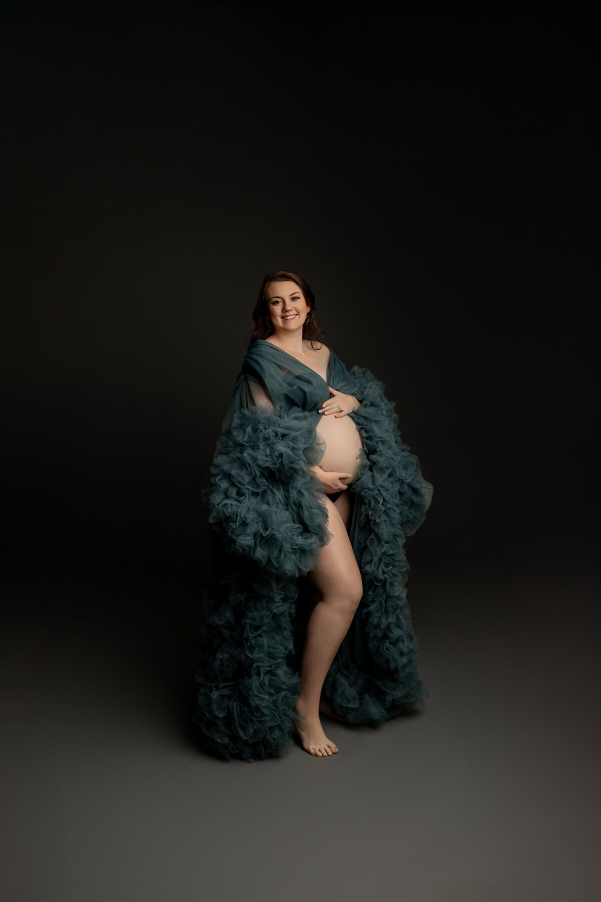 The expectant mother in green dress poses with a soft smile, gently cradling her belly as she stands gracefully in front of a dark backdrop. Captured by Inland Empire maternity photographer Kaitlyn Dawn Photography.