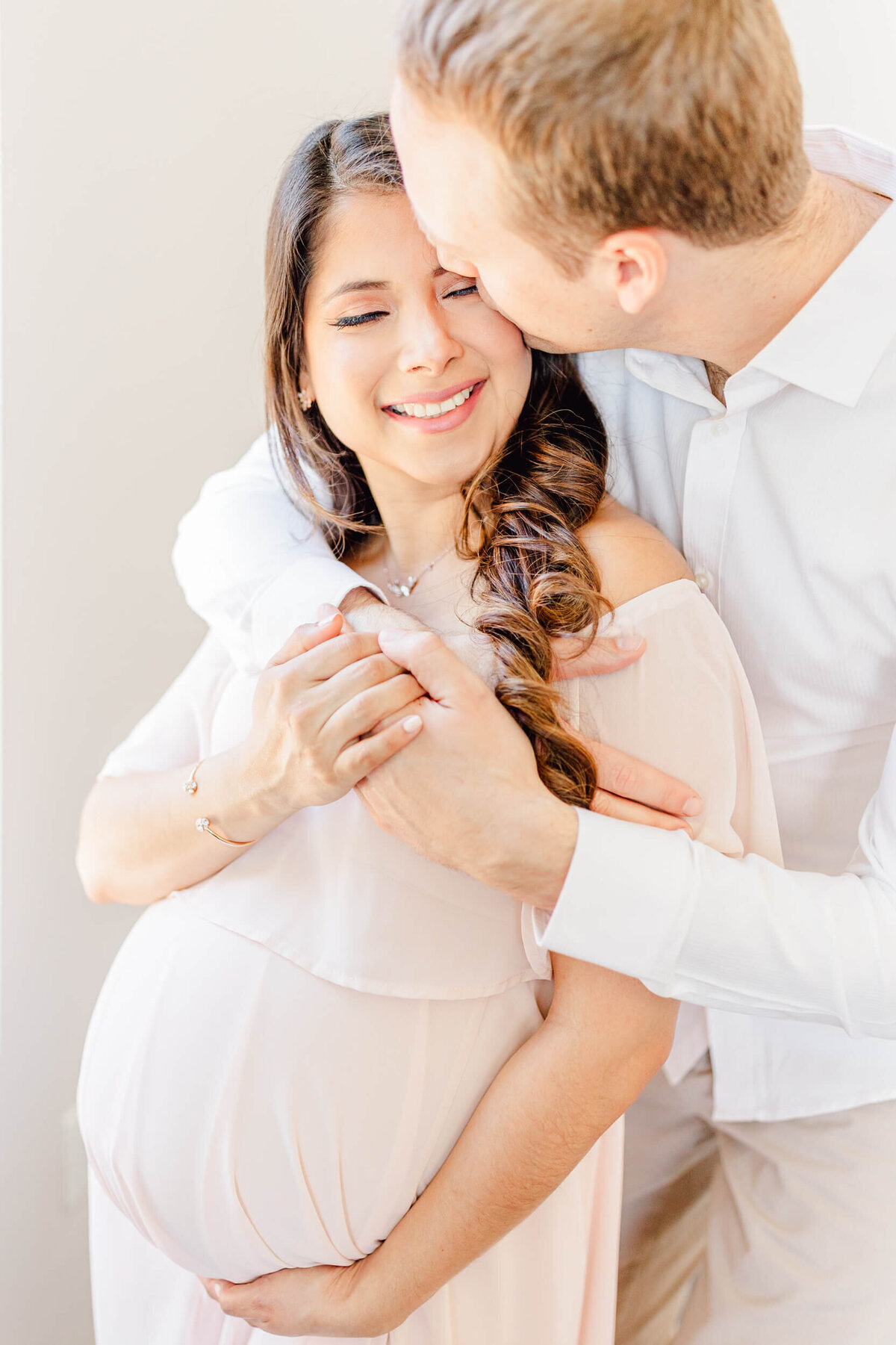 Pregnant woman in a blush pink dress smiles at her husband, who pulls her close to him