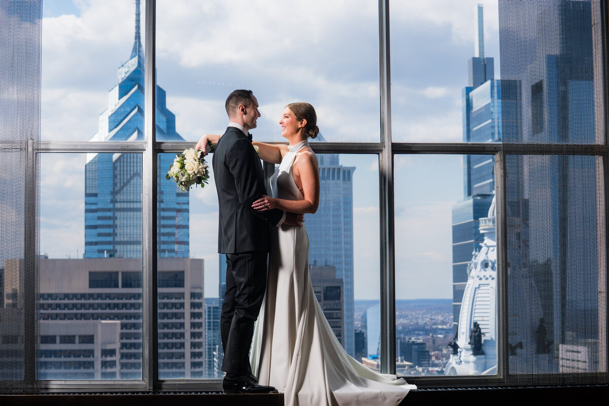 Bride and Groom standing on the windowsill of the loews hotel with the city in the back 