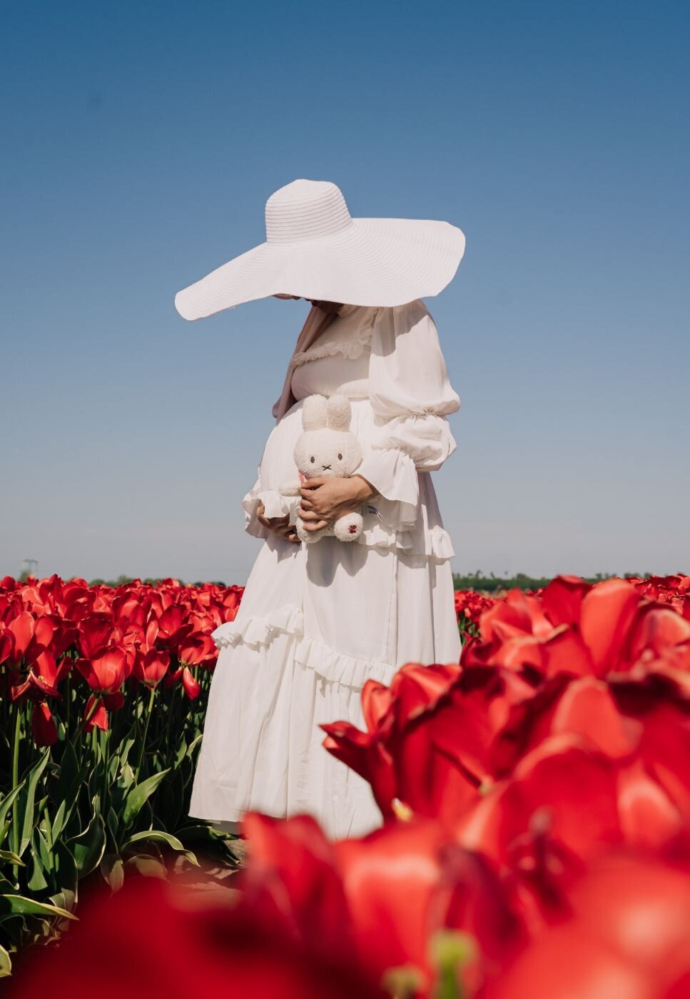 Pregnant woman in tulip field – Expectant mother in a flowing white dress and wide-brim hat holding a plush bunny among vibrant red tulips under a clear blue sky.