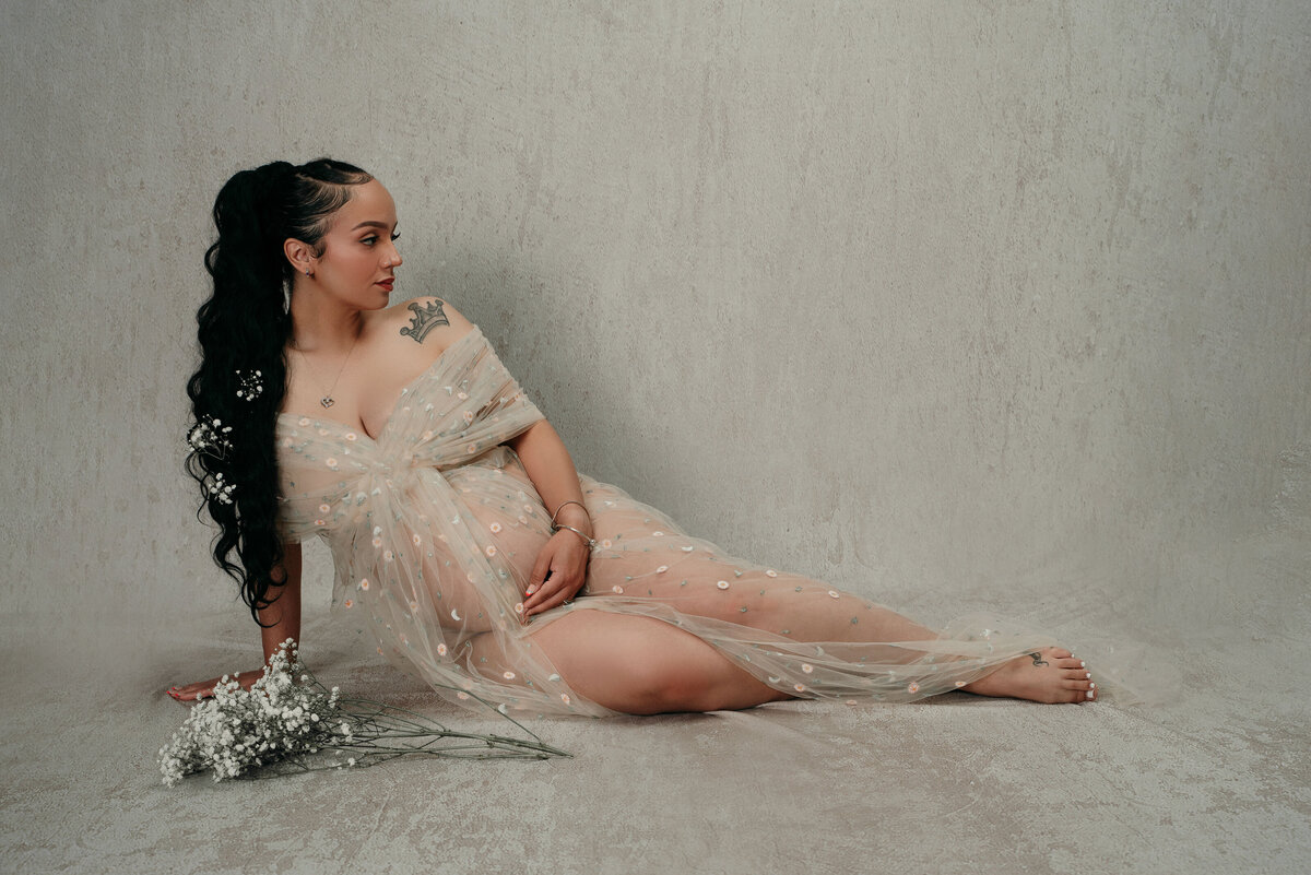 35 weeks pregnant woman with long black curly pony tail posing sitting down on light gray backdrop wearing grey tulle dress holding baby bump looking to the side