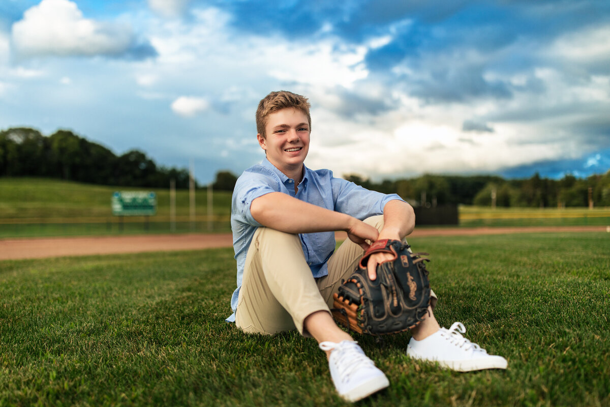 A red haired boy sits on a field that he plays at wearing a black baseball glove with a beautiful clouds behind him for his senior portraits