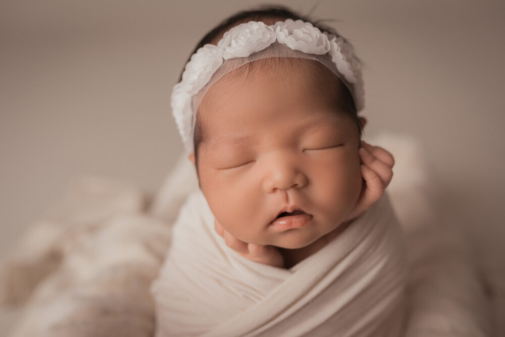 Atlanta, Georgia newborn photographer photographing newborn portrait session at atlanta ga maternity and newborn photography studio casey mcminn photography with baby girl in white flower headband, cream swaddle and hands on cheeks
