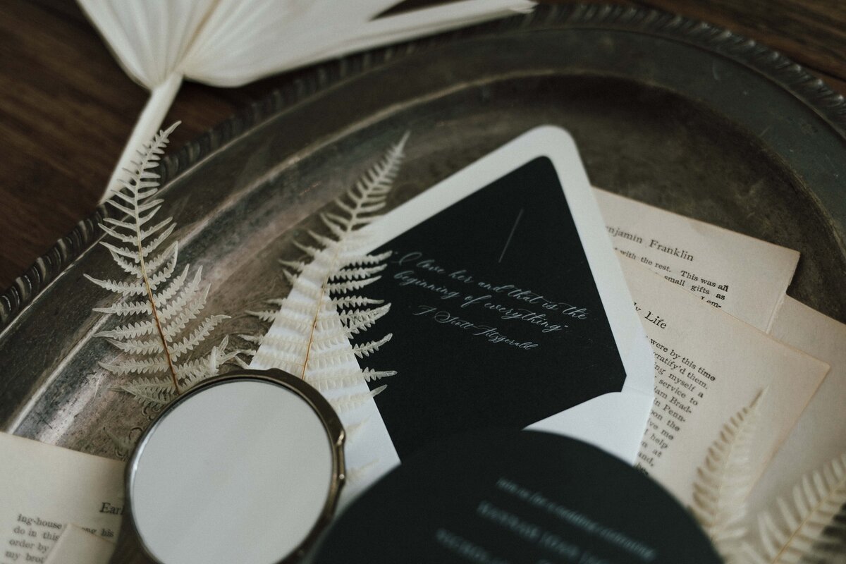 Mixed white and dark green wedding stationery with cursive white font atop a silver tray with loose book pages.