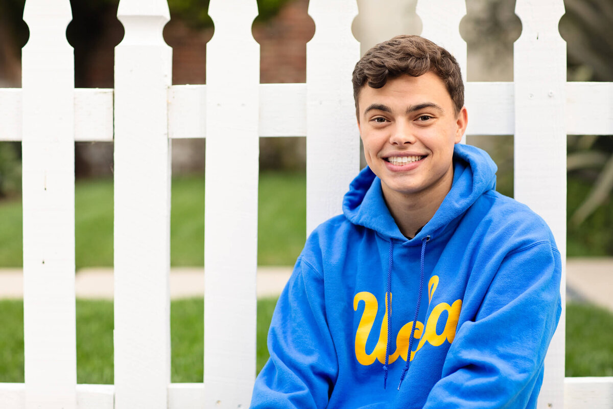 A young person in a blue UCLA hoodie sits in front of a white picket fence, smiling. The background shows green grass and blurred foliage.