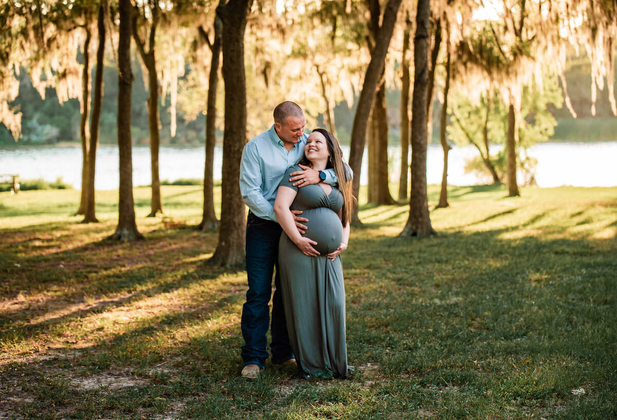 A husband hugs his wife from behind and smiles at her while she holds her baby bump.