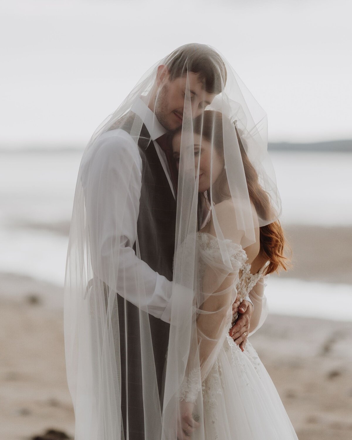 Couple have portraits on the beach in Tenby, with a veil draped over the top of them.