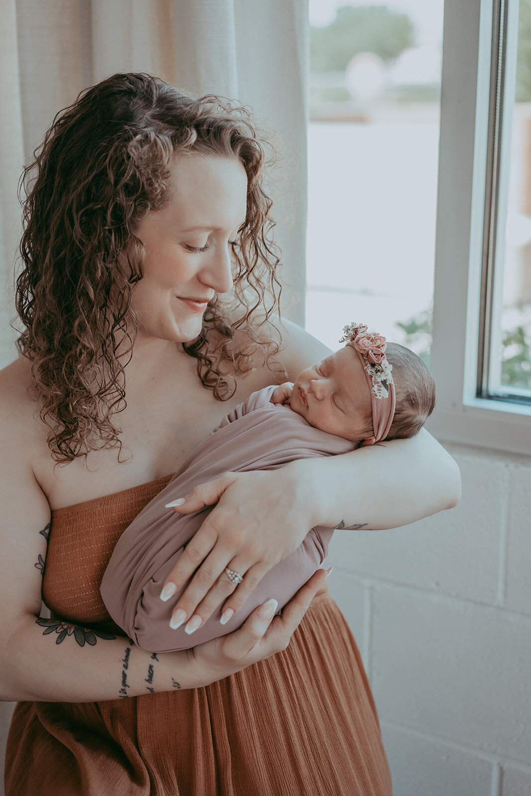 mom sits next to bright window adoring her newborn daughter swaddled in mauve