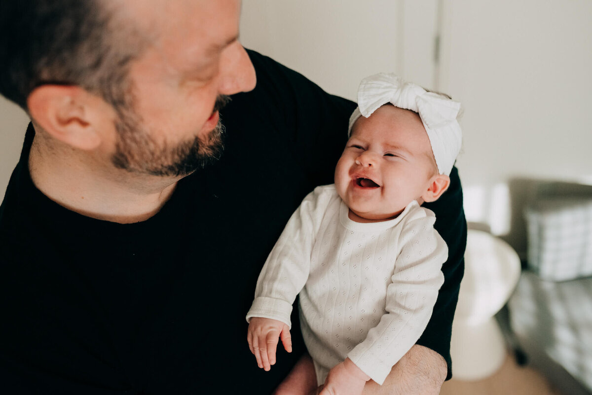 A baby girl wearing a white bow gives her father a bright smile as he holds her in his arms during their photoshoot with Allison Wolf, Boston Newborn Photographer
