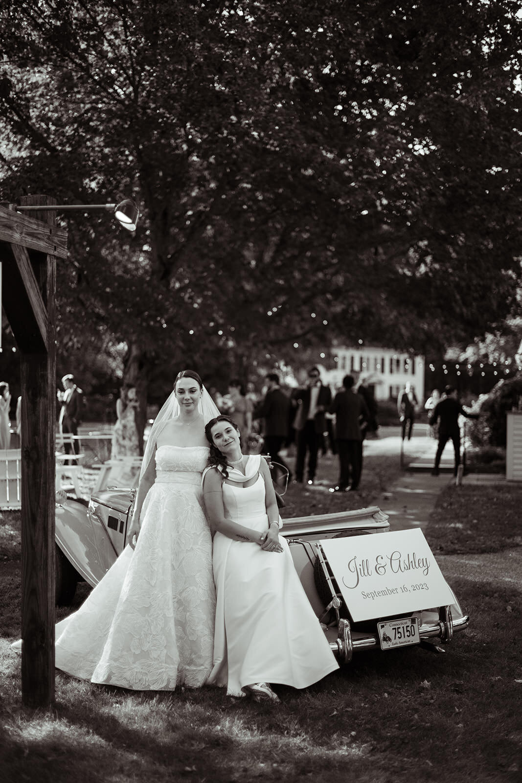 Two brides leaning against vintage car with "Jill & Ashley" sign on the back