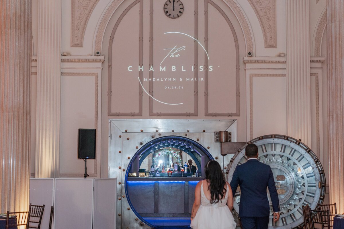 A bride and groom make their grand reception entrance into the iconic vault bar at The Treasury on the Plaza in St. Augustine, Florida. The venue’s dramatic architecture and custom monogram projection create a modern, luxurious atmosphere. The couple walks hand in hand toward the glowing bar, ready to celebrate with their guests. Captured by Phavy Photography in a true-to-color, documentary style that blends elegance, storytelling, and the vibrant energy of a St. Augustine wedding.
