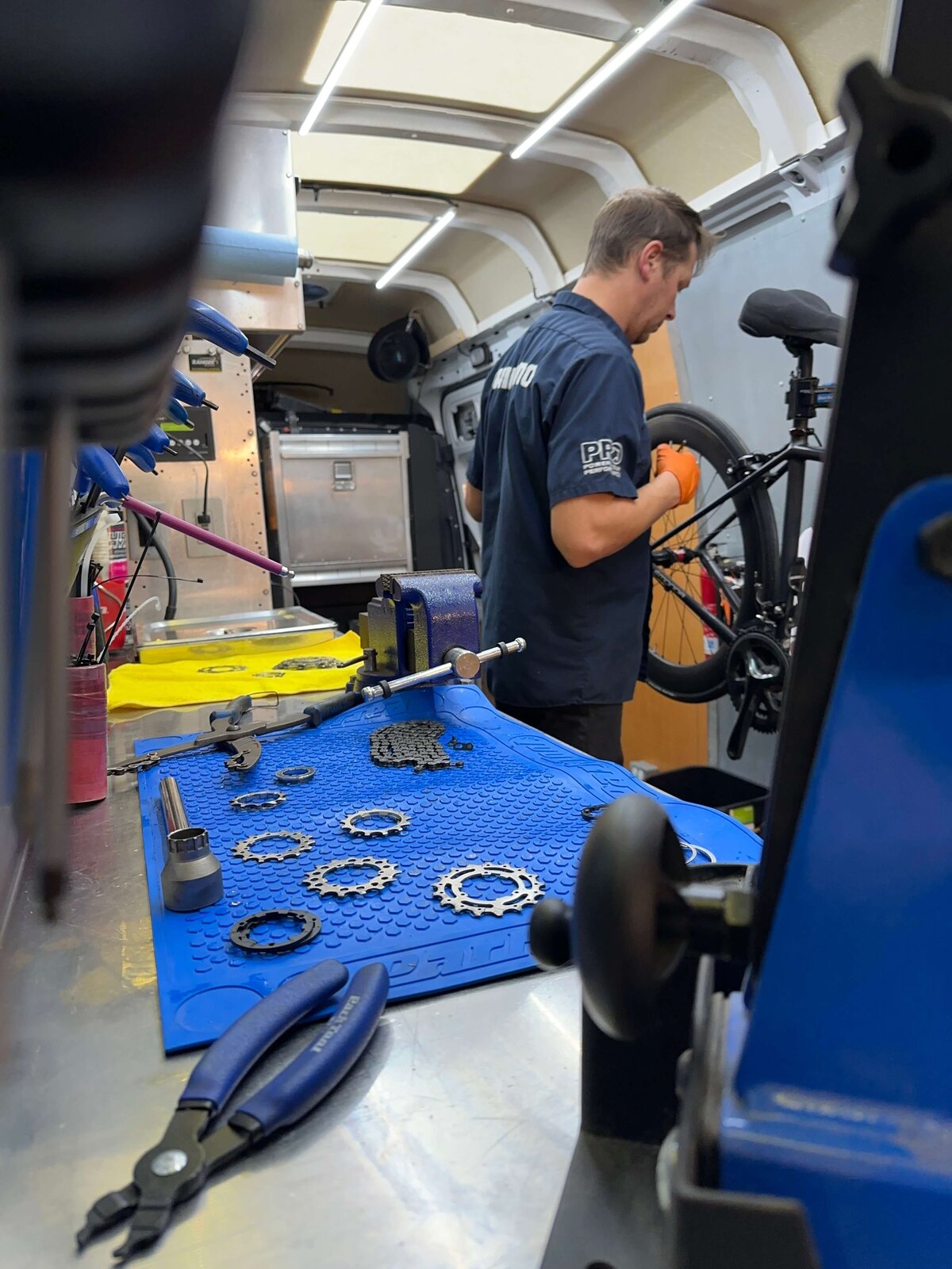 A person wearing a black shirt and orange gloves works on a bicycle inside a mobile workshop. Various tools and bike parts are arranged neatly on a blue mat on the workbench. The workshop interior is well-organized and brightly lit