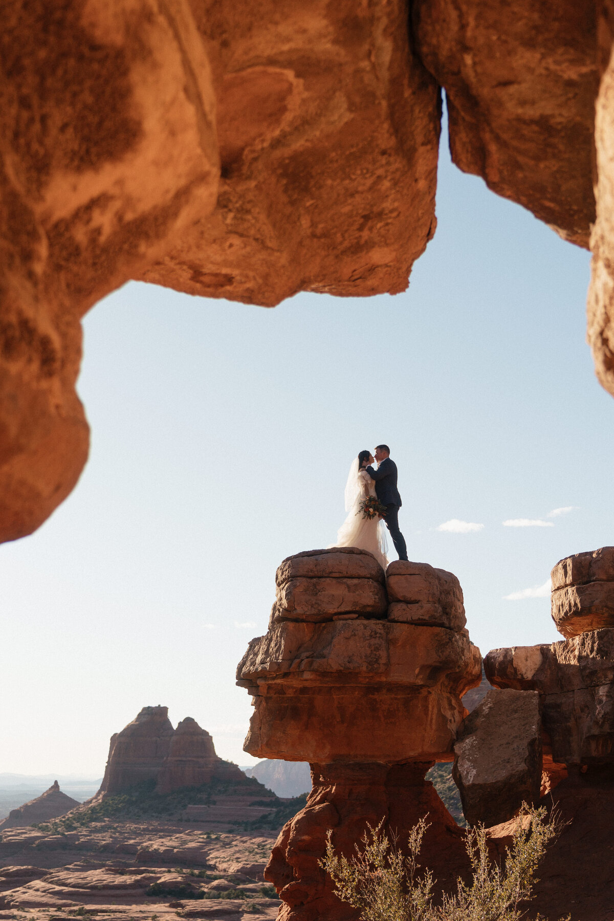 Bride and groom standing on a dramatic red rock ledge at Merry Go Round Rock in Sedona during their adventurous elopement, framed by towering sandstone formations and sweeping desert views.