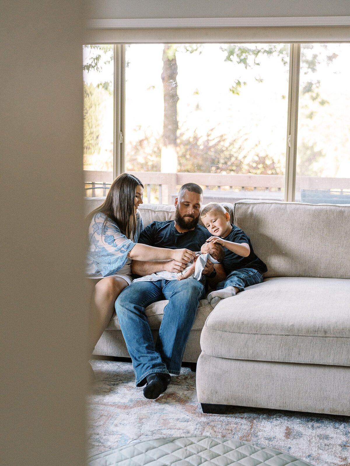 A family sitting on a couch in front of a window, smiling at a baby in their arms.