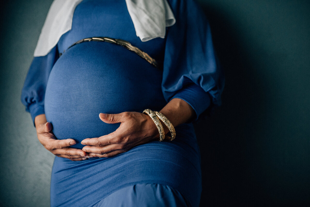 Pregnancy photoshoot with woman wearing blue dress, white scarf, and holding her belly.