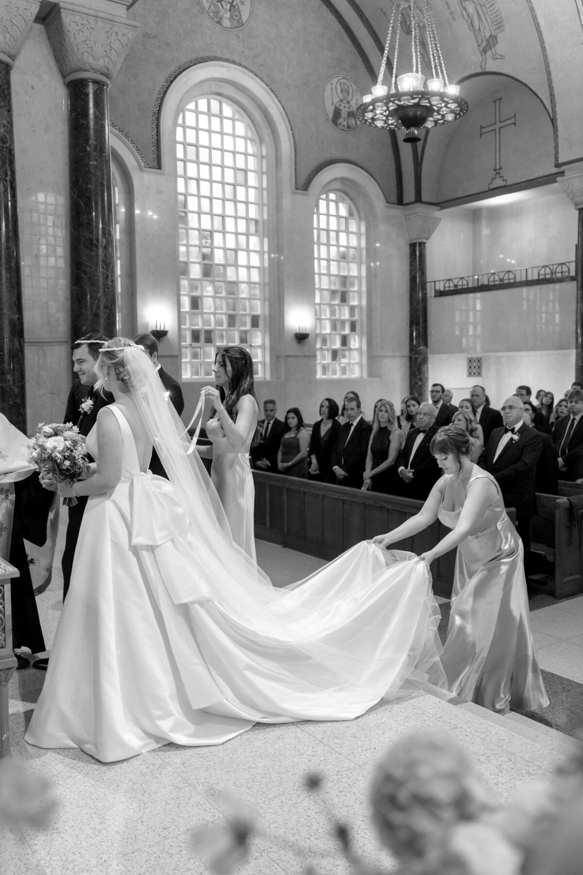 A black and white photo of a bride in a long gown and veil standing at the altar in a church. Bridesmaids adjust her train. Guests are seated, and the churchs arched windows and ornate ceiling are visible.
