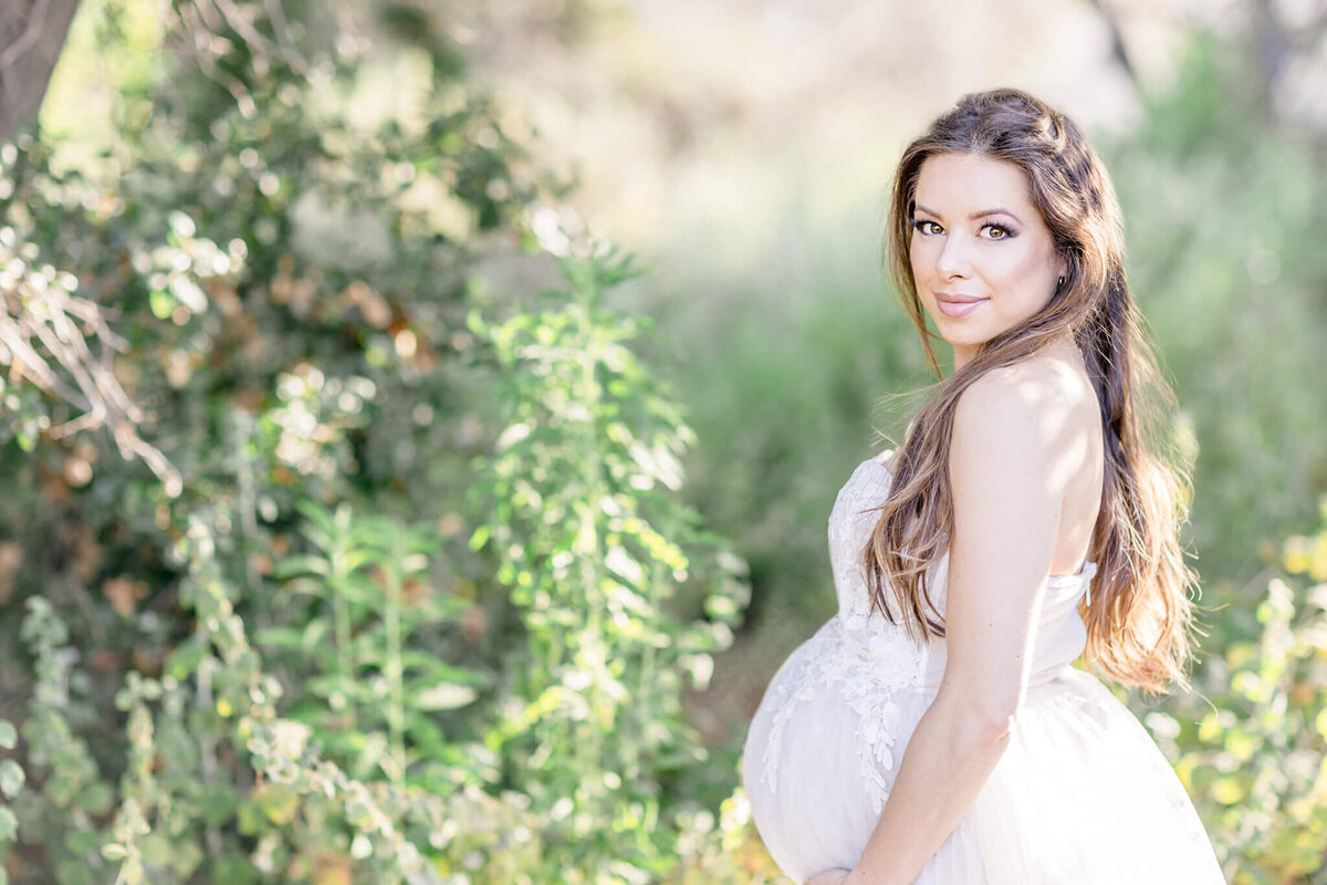 A pregnant woman in a white dress stands outdoors, surrounded by vibrant greenery. She has long, wavy hair and is looking over her shoulder with a serene expression. The sunlight creates a soft, natural atmosphere.
