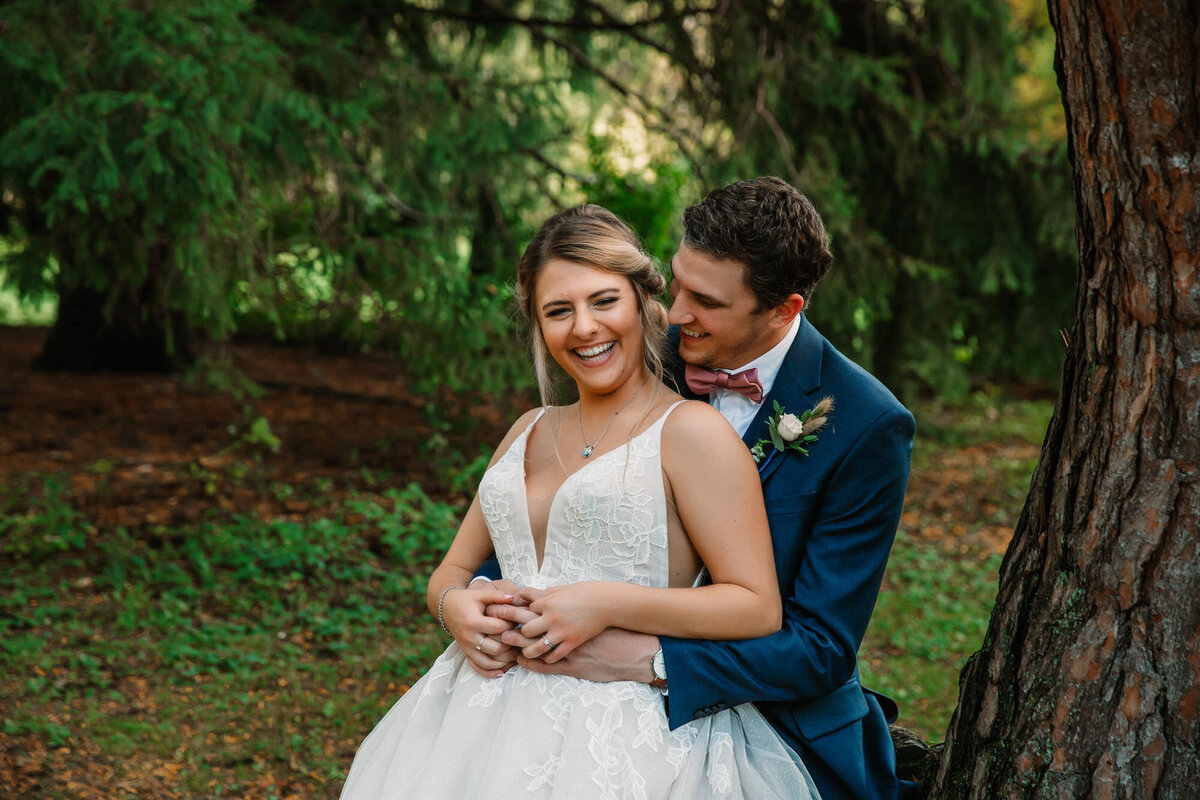 groom holds bride from behind while they laugh sitting on a tree