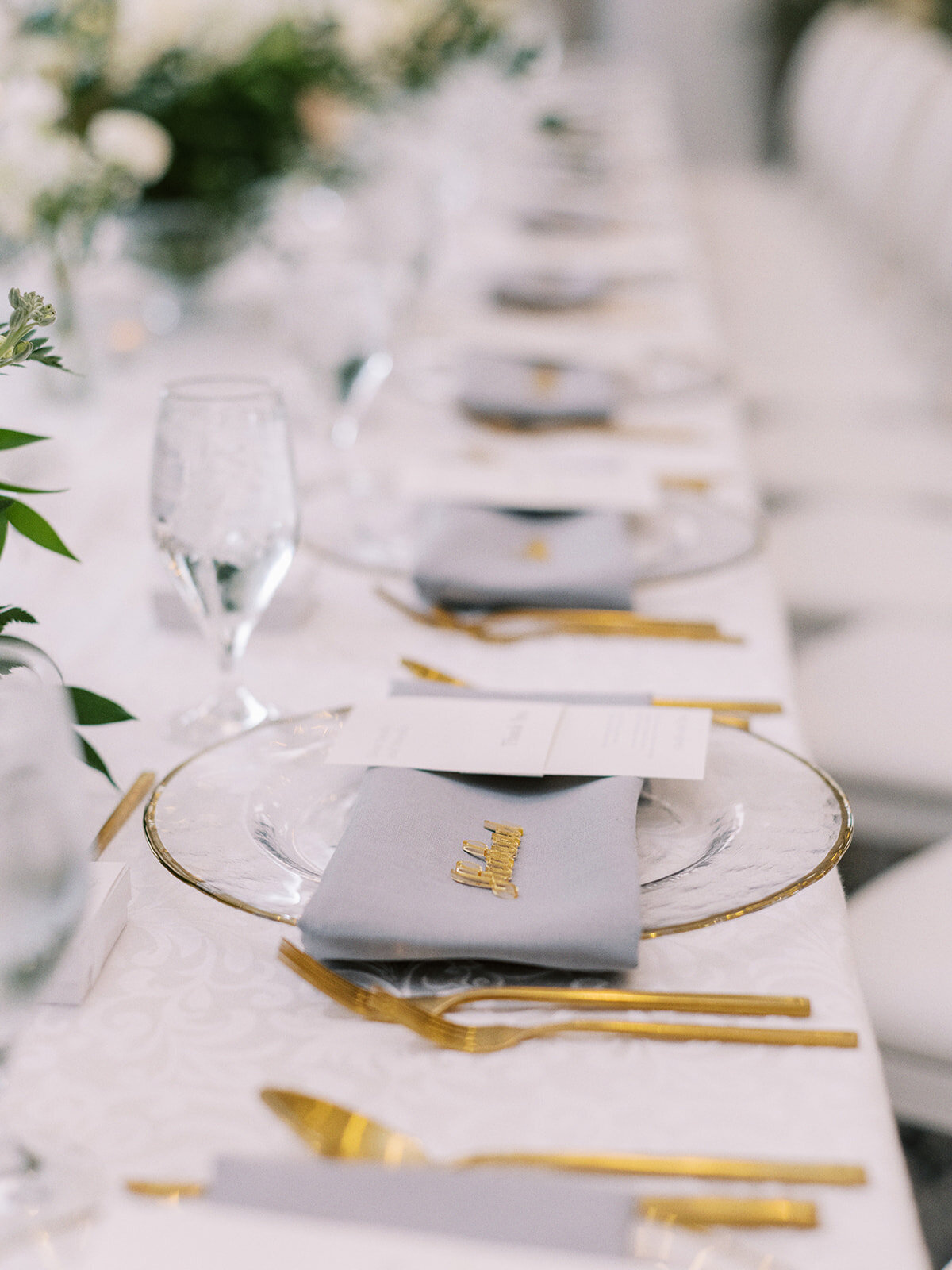 A table set with gold cutlery, clear glasses, white plates, and grey napkins arranged neatly, each with a menu card placed on top. The table is adorned with greenery and white floral arrangements, perfect for a classic Calgary wedding at the Fairmont Palliser.