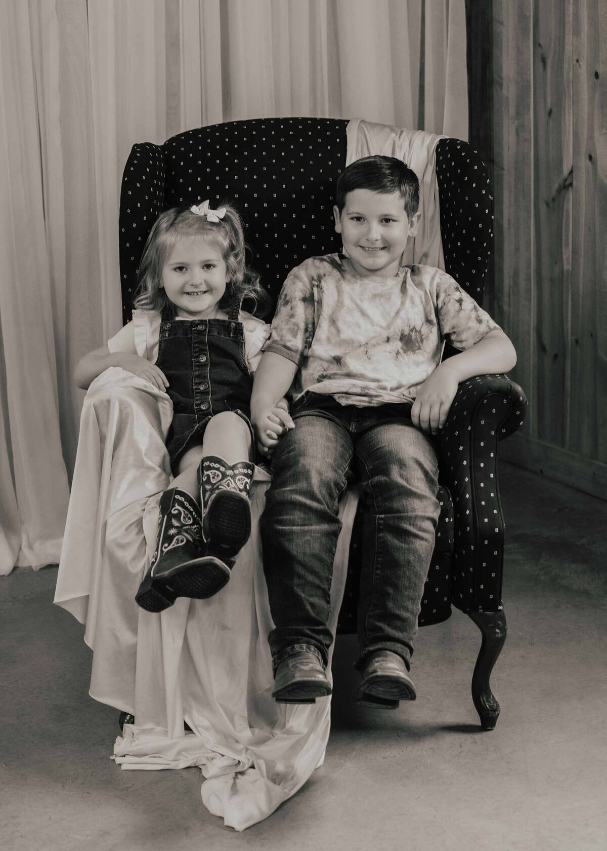 Maddie Rae Photography black and white picture of two little kids, brother and sister, sitting in a chair and holding hands, they are smiling at the camera