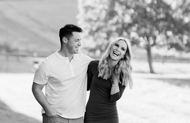A smiling couple enjoys a sunny park in this black and white image, embodying the essence of New England elopement photography. The man, in a light shirt and pants, lovingly wraps his arm around the woman in a dark dress. Trees and grass bask in the backdrop of their joyful moment.